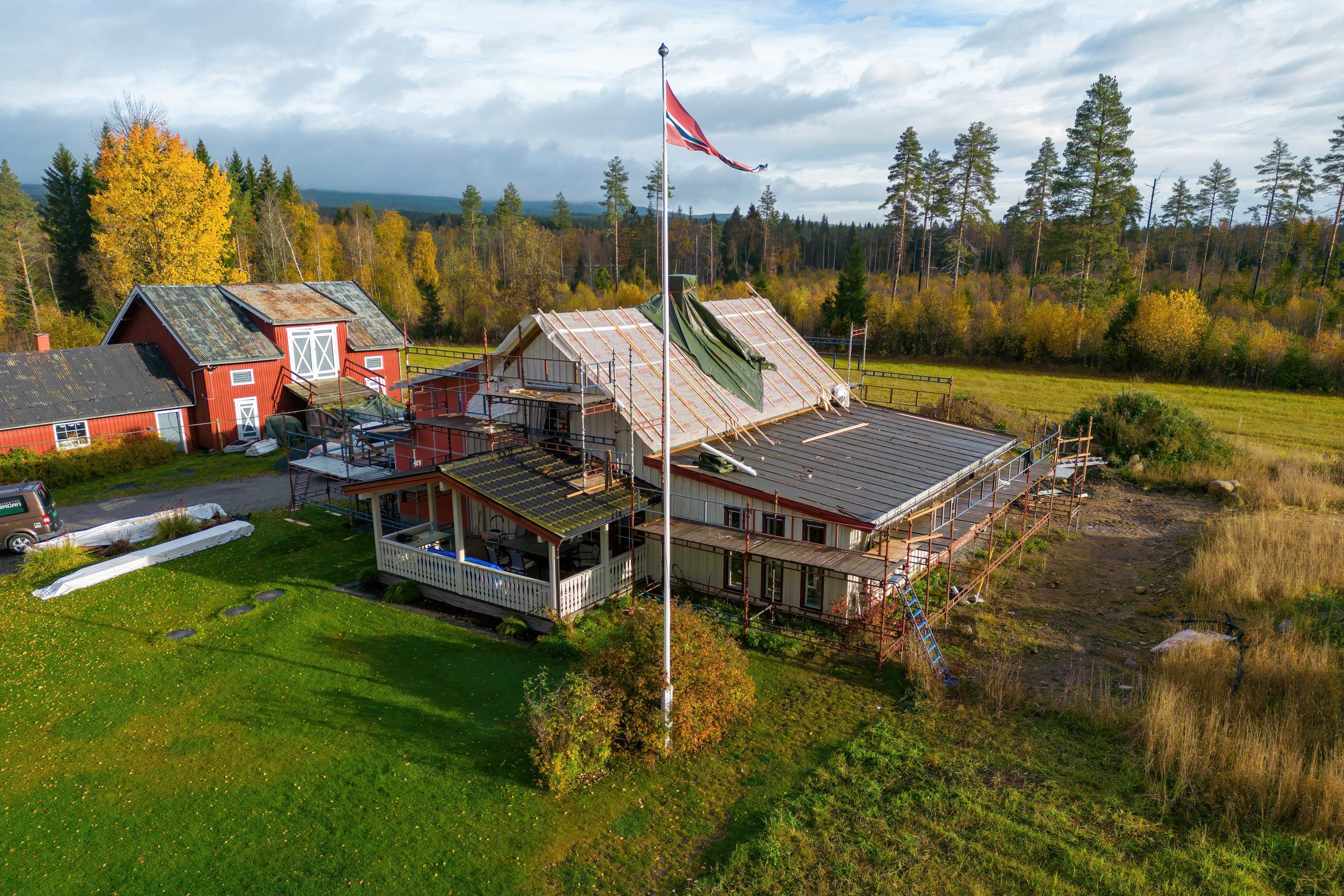 Dronebilde av et pågående rehabiliteringsprosjekt: hovedhus med stillaser rundt taket, sentralt flaggstang med norsk flagg, rød låve til venstre og høstskog i bakgrunnen.